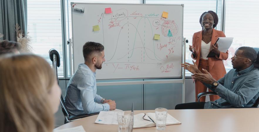 People sitting around a board table with computers and papers looking at whiteboard with woman standing next to white board.