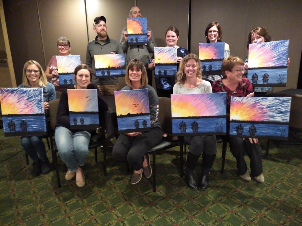 A group of people, one row sitting and one row standing, holding their finished sunset paintings. The paints are all of the same scene, but each person has painted it slightly differently.